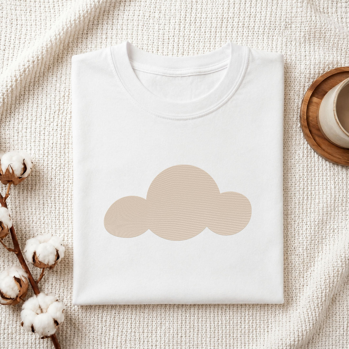 Folded white cotton t-shirt mockup featuring a minimalist cloud machine embroidery design, styled with cotton stems and a wooden cup   