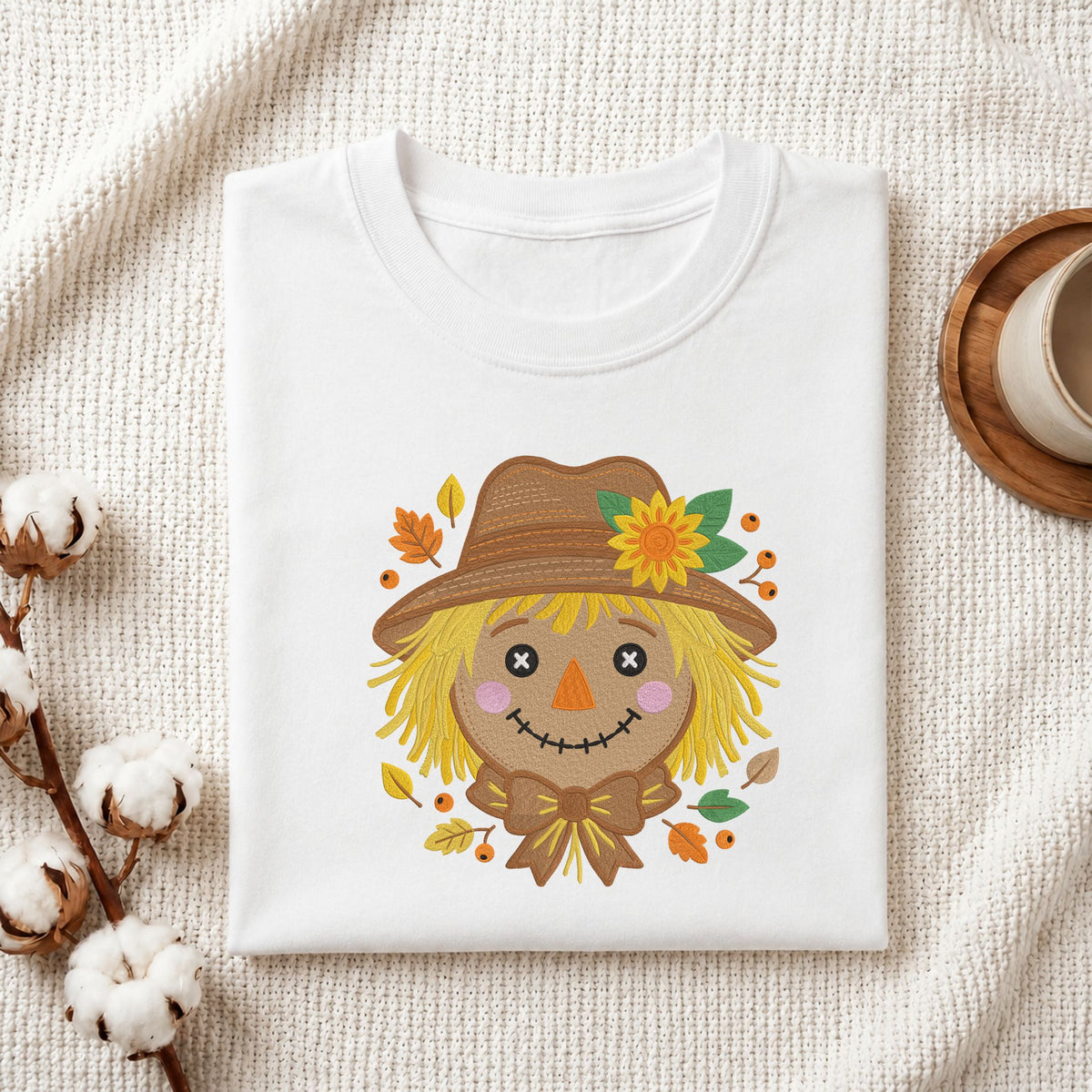  Folded white cotton t-shirt mockup featuring a cute autumn scarecrow face machine embroidery design, styled with cotton stems and a wooden cup   