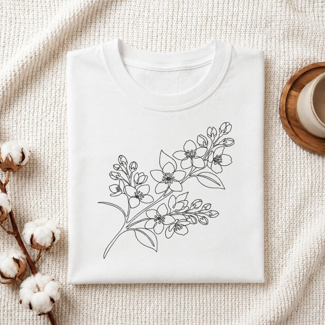 A folded white crewneck t-shirt with a black line art floral branch design on the front, styled on a textured cream knit blanket with cotton bolls and a wooden cup in the background.