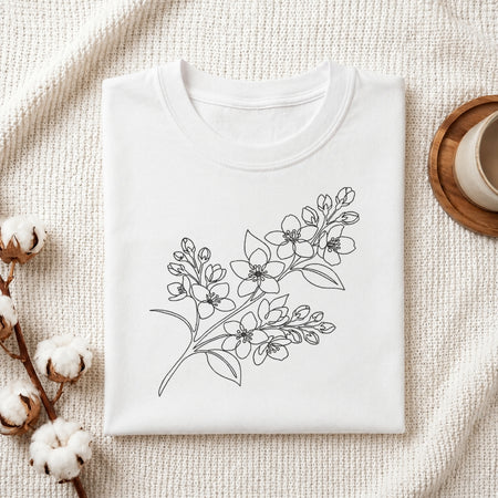 A folded white crewneck t-shirt with a black line art floral branch design on the front, styled on a textured cream knit blanket with cotton bolls and a wooden cup in the background.