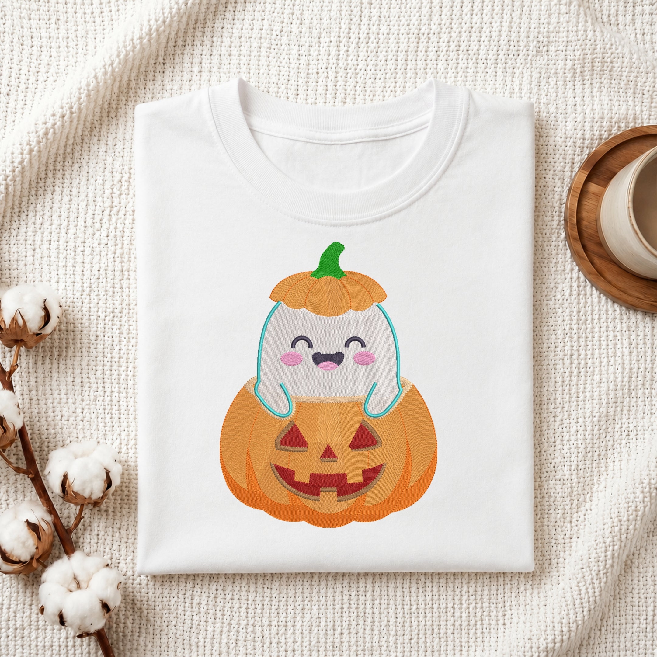 A folded white crewneck t-shirt featuring a cute cartoon graphic of a smiling ghost inside a jack-o'-lantern pumpkin, styled on a textured cream fabric with cotton stems and a wooden cup.