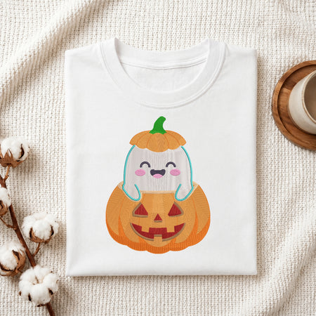 A folded white crewneck t-shirt featuring a cute cartoon graphic of a smiling ghost inside a jack-o'-lantern pumpkin, styled on a textured cream fabric with cotton stems and a wooden cup.