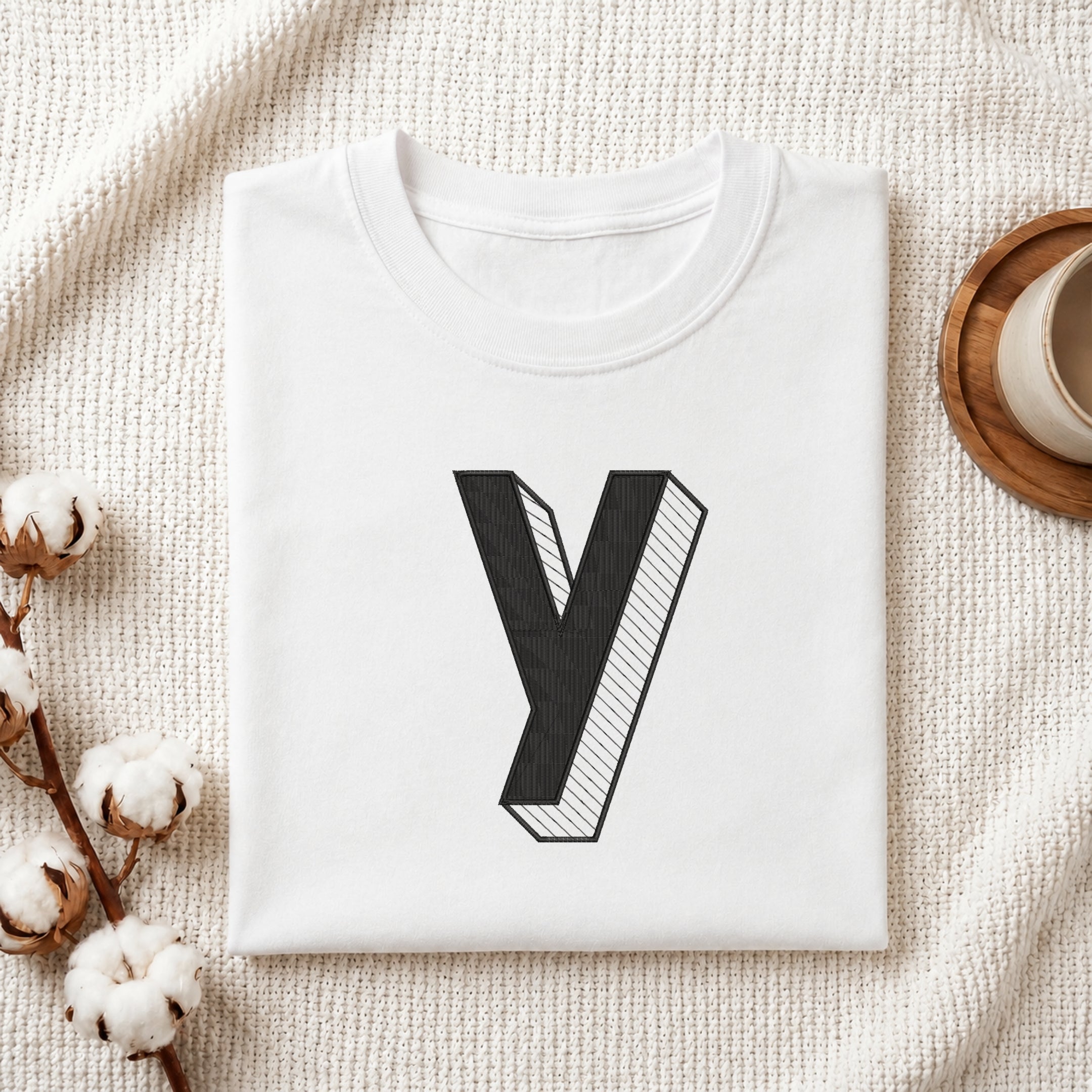 A folded white t-shirt with a large black 3D-style letter 'y' graphic on the front, displayed on a textured cream fabric background with cotton stems and a cup on a wooden coaster.