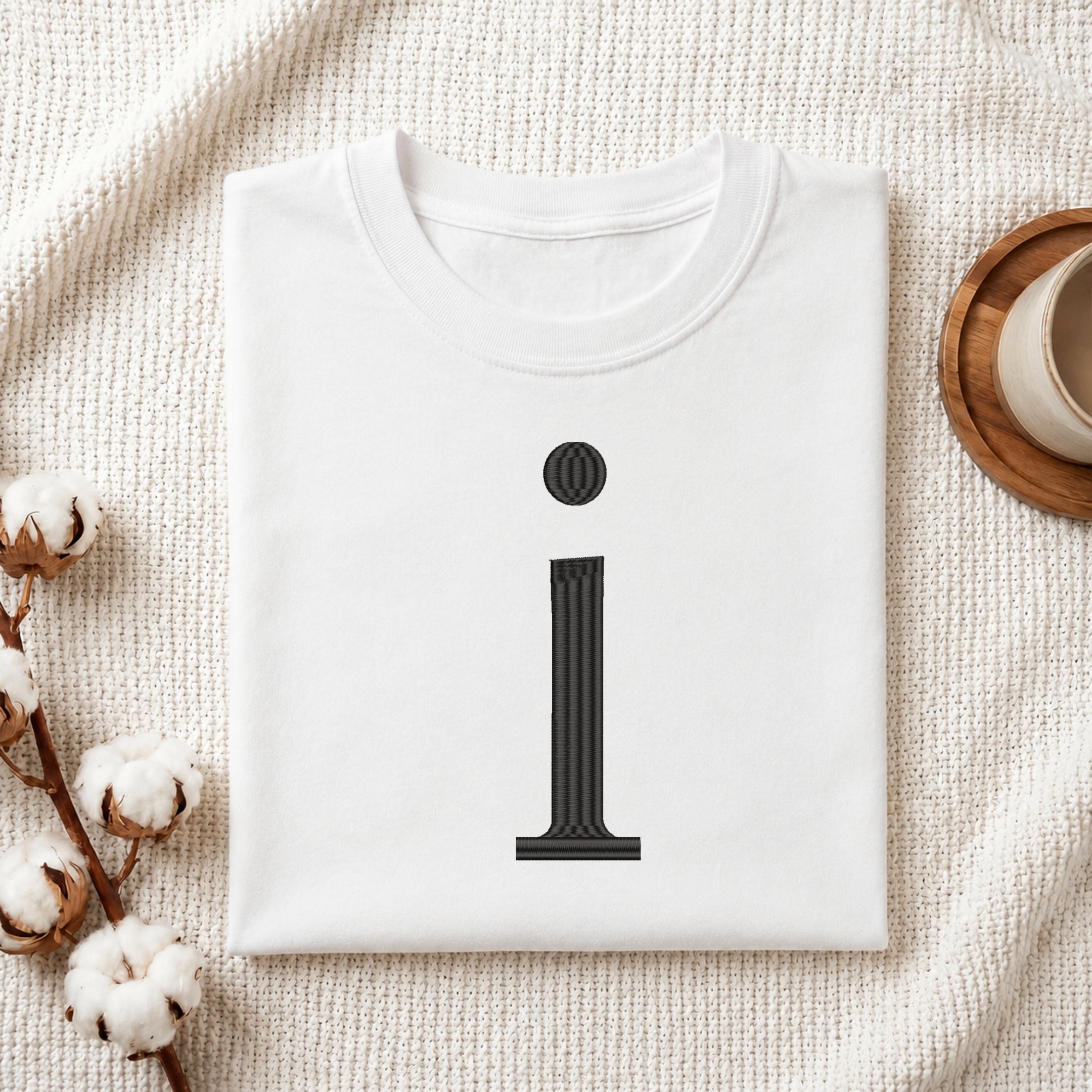 A folded white crewneck t-shirt with a black graphic of the letter 'i' styled like a classical column, presented in a flat-lay setting on a textured cream fabric with cotton stems and a cup in the background.