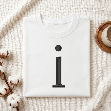 A folded white crewneck t-shirt with a black graphic of the letter 'i' styled like a classical column, presented in a flat-lay setting on a textured cream fabric with cotton stems and a cup in the background.