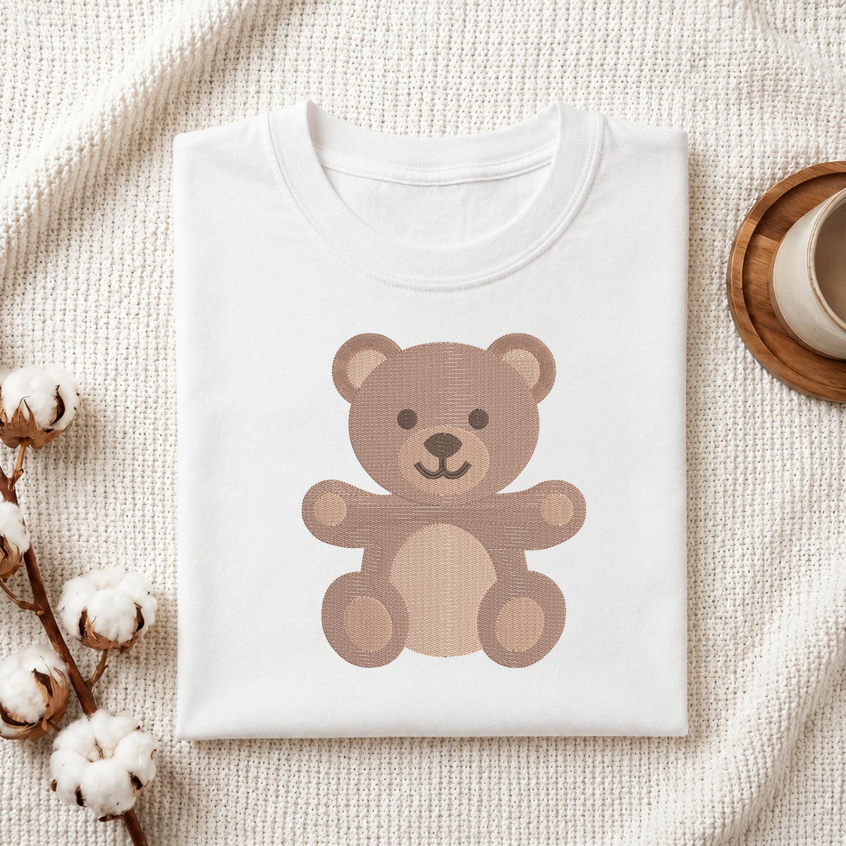 A folded white T-shirt with a cute cartoon teddy bear graphic in shades of brown, styled on a textured cream fabric with cotton stems and a wooden cup in the background.