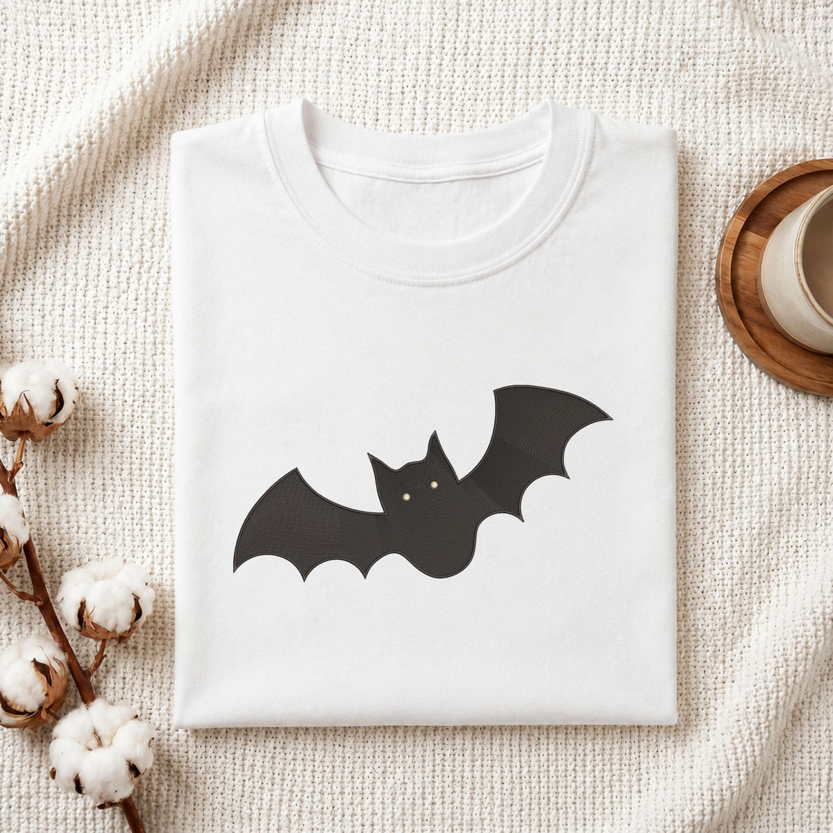 Folded white cotton t-shirt mockup featuring a spooky cute black bat machine embroidery design, styled with cotton stems and a wooden cup.