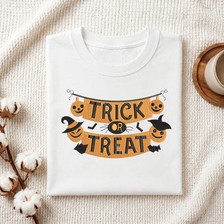 A folded white T-shirt with a Halloween-themed 'Trick or Treat' banner graphic, styled on a textured cream fabric with cotton stems and a small cup in the corner.