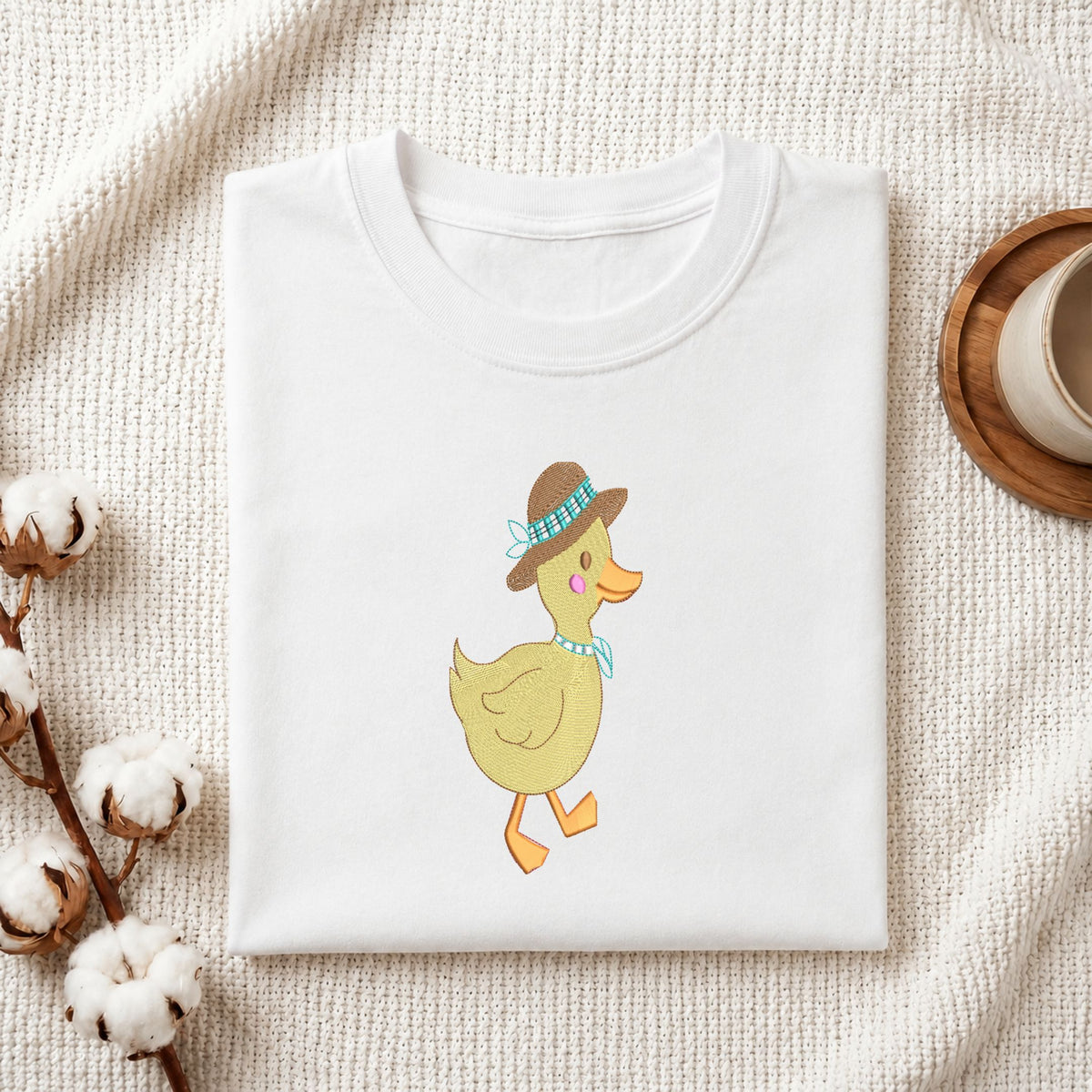 Folded white cotton t-shirt mockup featuring a cute duck in a hat machine embroidery design, styled with cotton stems and a wooden cup.