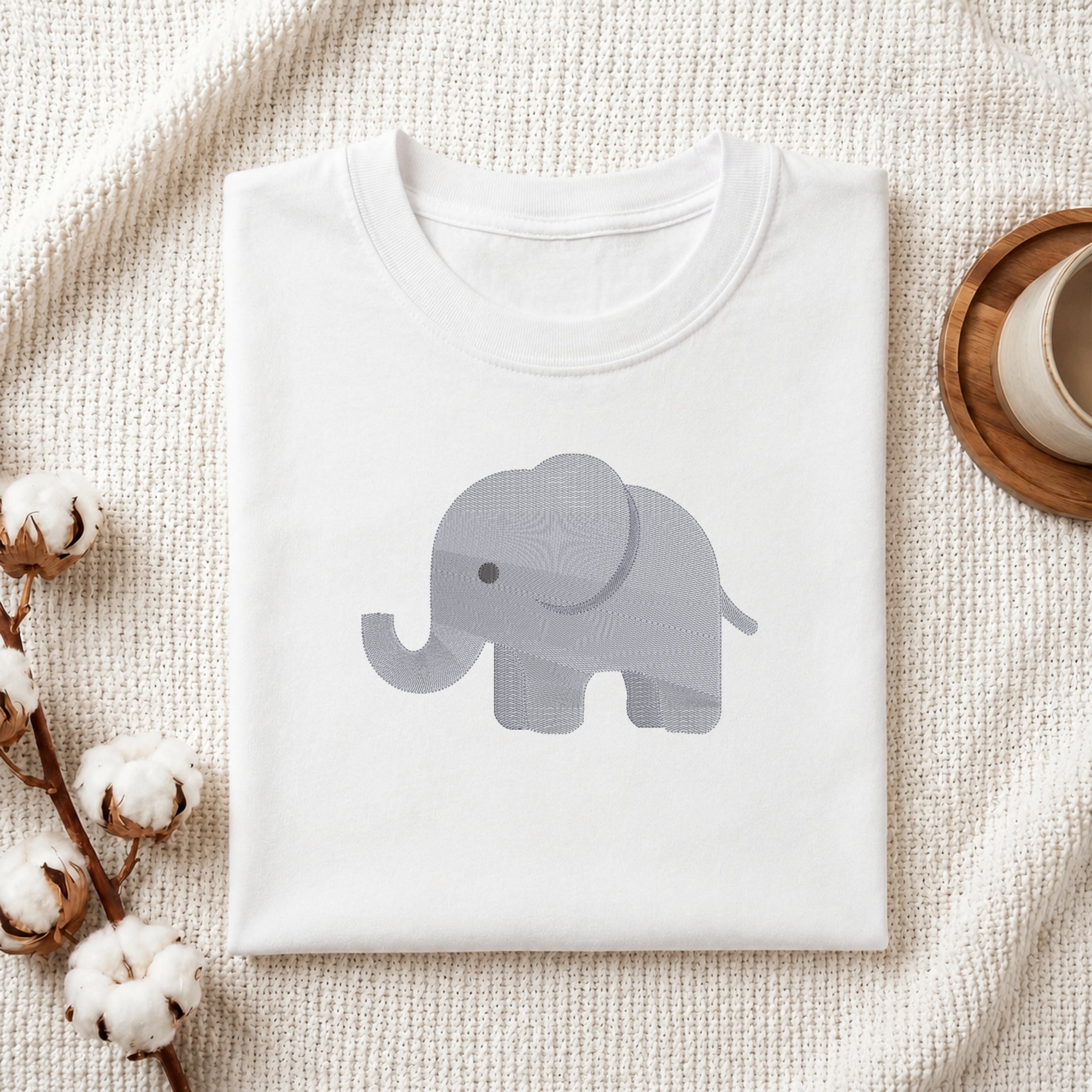 A folded white crewneck t-shirt featuring a light gray patterned elephant graphic on the chest, styled on a textured cream knit blanket with cotton bolls and a wooden cup in the background.