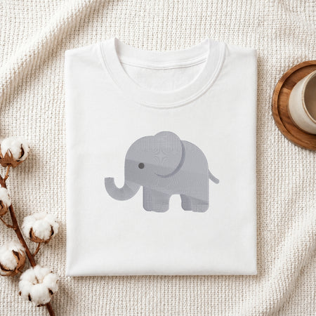 A folded white crewneck t-shirt featuring a light gray patterned elephant graphic on the chest, styled on a textured cream knit blanket with cotton bolls and a wooden cup in the background.
