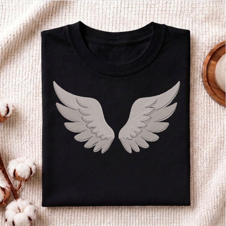 A folded black crewneck t-shirt with a large gray embroidered angel wings graphic on the front, displayed on a textured beige fabric background with cotton bolls and a wooden tray.