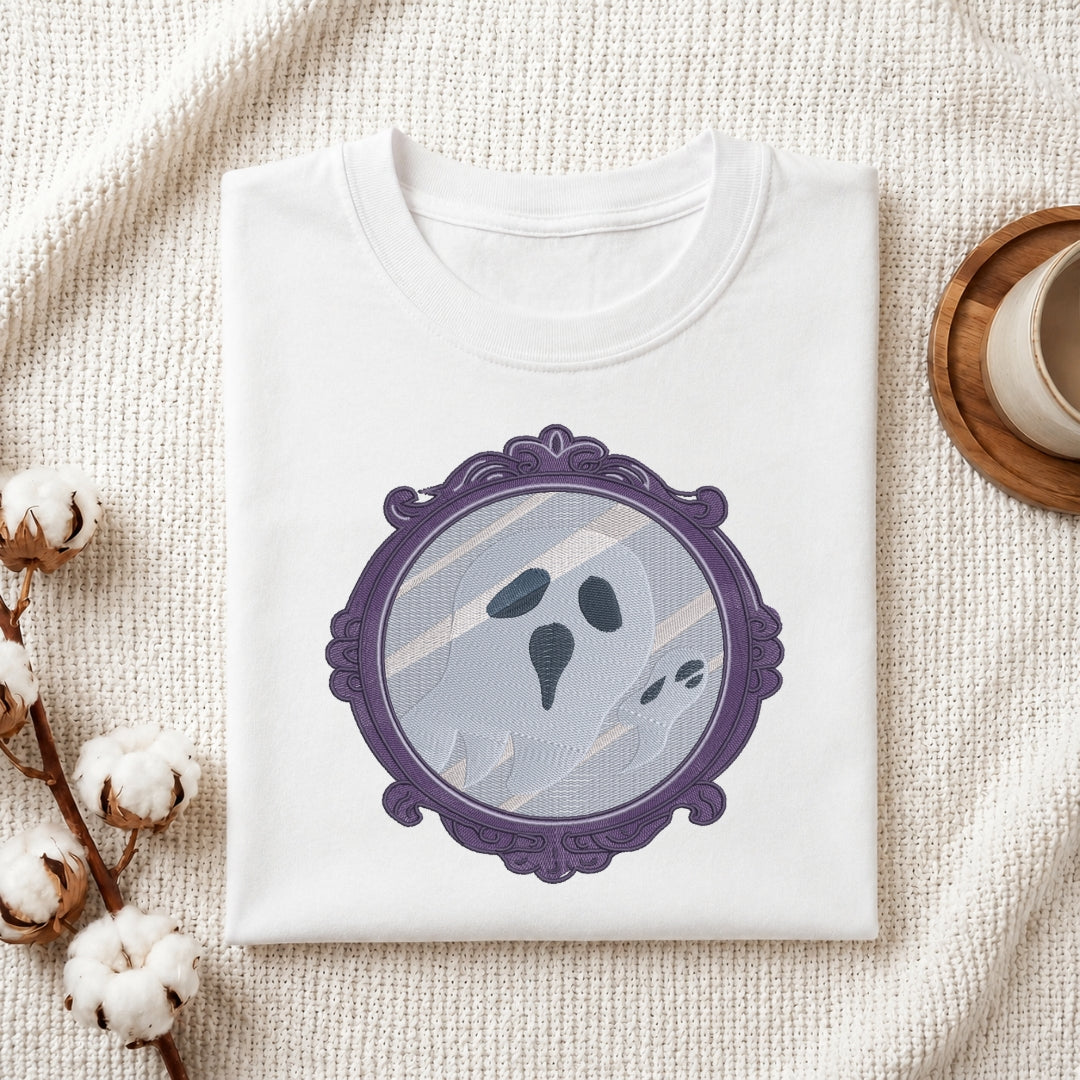 A folded white crewneck t-shirt with a graphic of a ghostly face inside an ornate purple mirror frame, styled on a textured cream fabric with cotton bolls and a wooden cup.