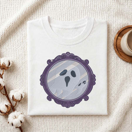 A folded white crewneck t-shirt with a graphic of a ghostly face inside an ornate purple mirror frame, styled on a textured cream fabric with cotton bolls and a wooden cup.