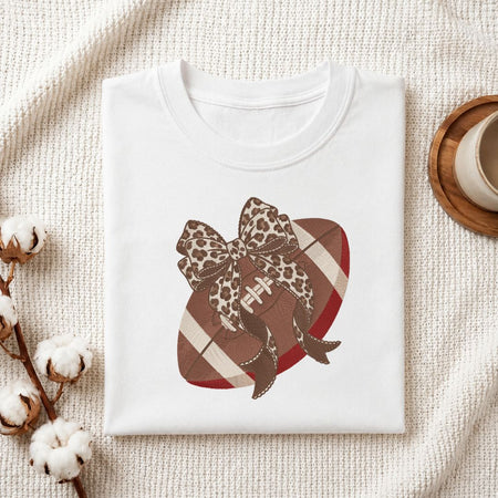 A folded white crewneck t-shirt displayed on a textured cream fabric background, decorated with cotton stems and a wooden cup. The t-shirt features a graphic of a football with red and white stripes, brown leopard print accents, and a large leopard print bow.