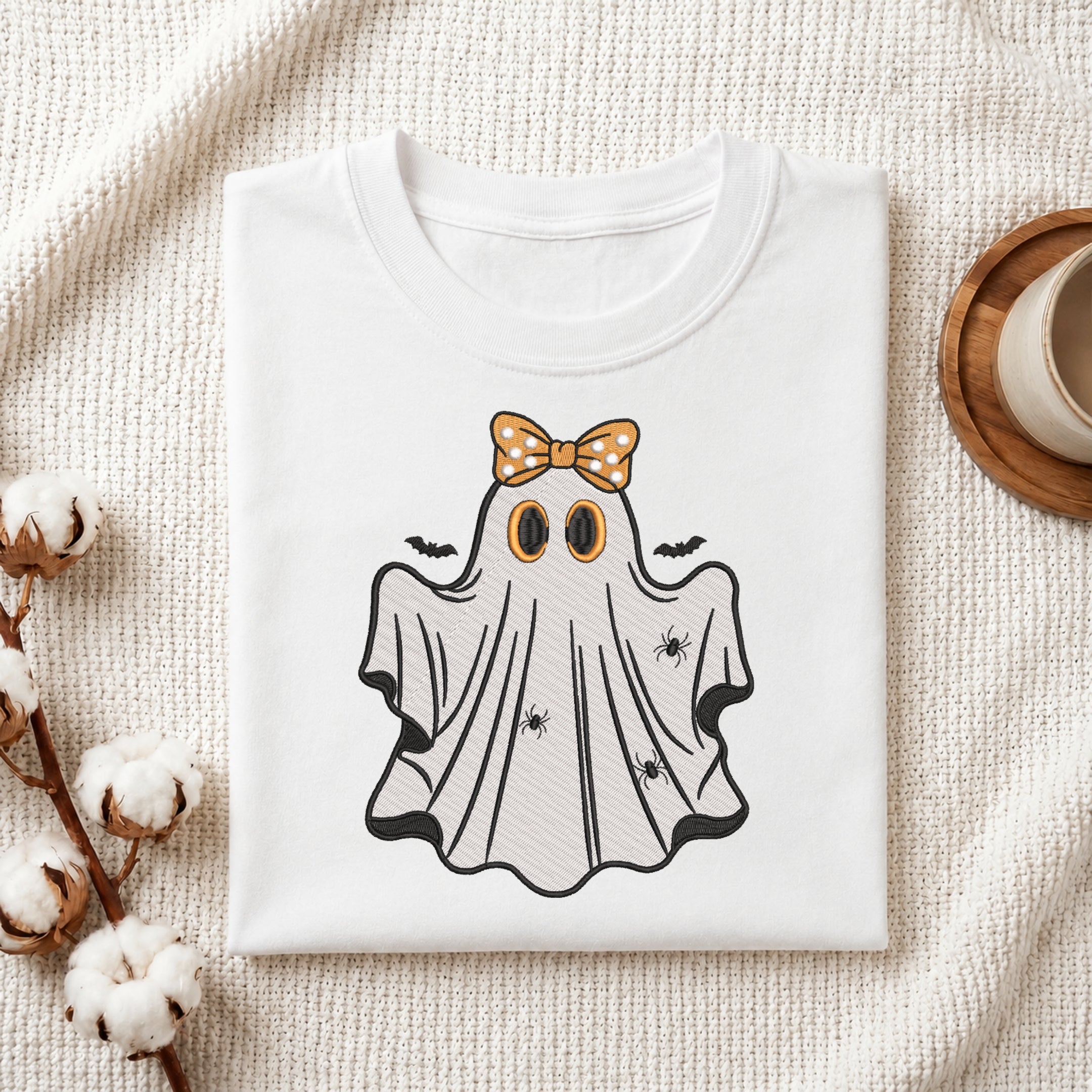 Folded white t-shirt featuring an embroidered ghost with an orange bow and tiny spiders.
