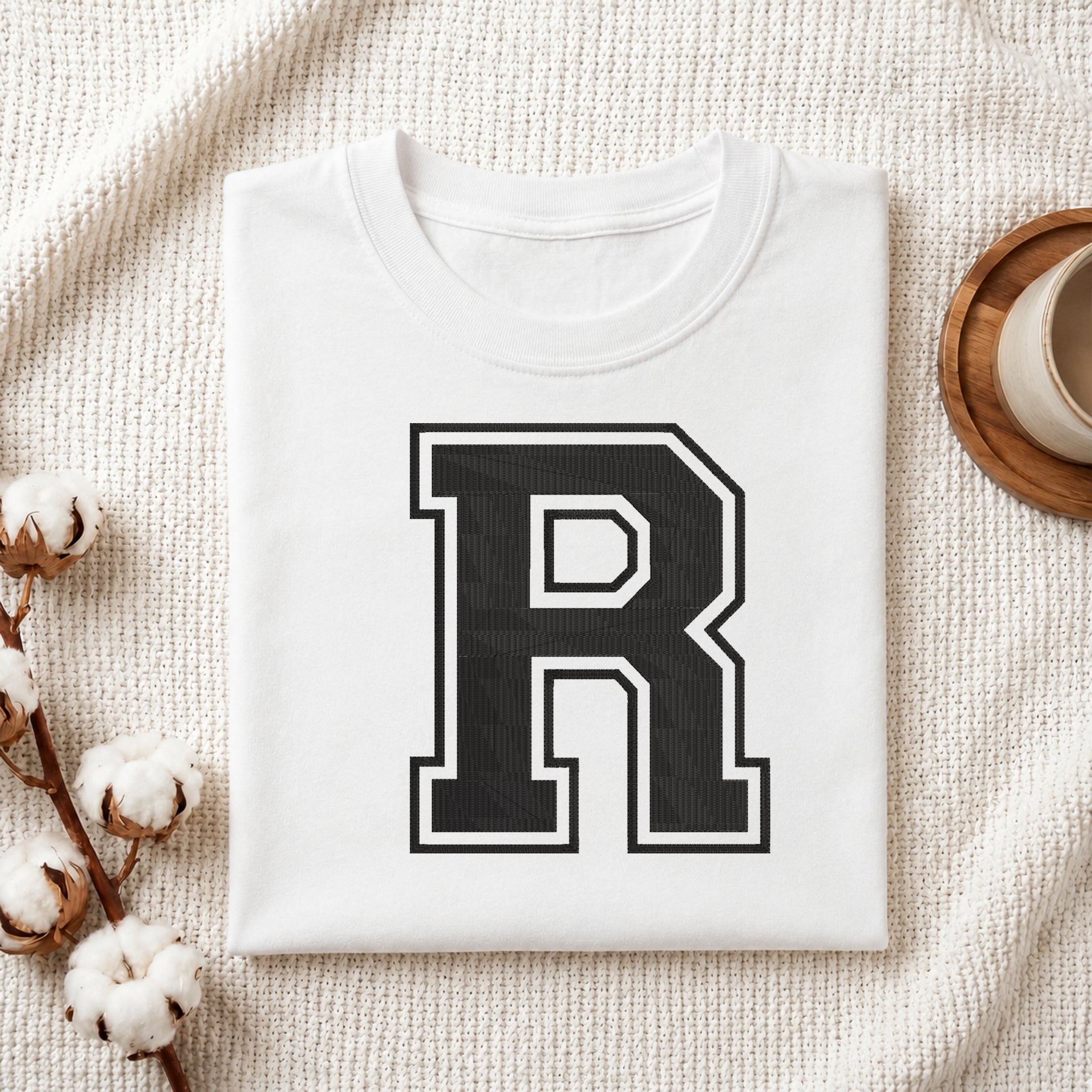 Flat-lay mockup showing a folded white t-shirt featuring a large collegiate-style embroidered black letter 'R' with a white border outline, set against a cozy white knitted blanket background.