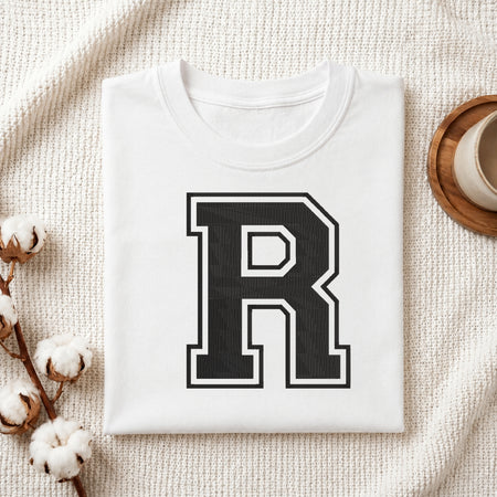 Flat-lay mockup showing a folded white t-shirt featuring a large collegiate-style embroidered black letter 'R' with a white border outline, set against a cozy white knitted blanket background.