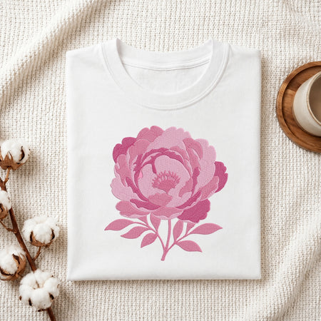 A folded white crewneck t-shirt with a large pink peony graphic on the front, styled on a textured cream knit blanket with cotton bolls and a wooden cup in the background.