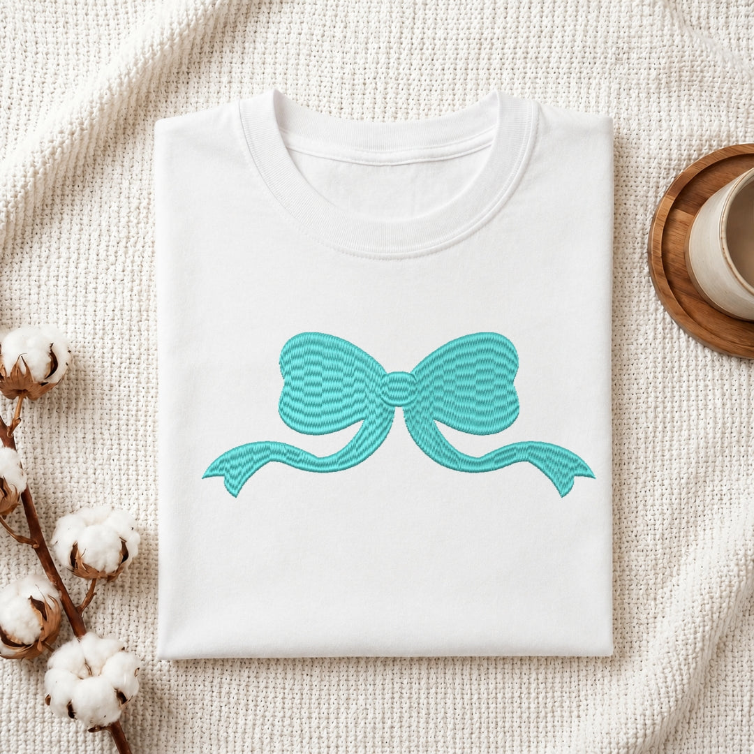 A folded white crewneck t-shirt with a turquoise bow graphic on the front, styled on a textured cream knit blanket with cotton bolls and a wooden cup in the background.