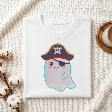 A folded white crewneck t-shirt featuring a cute cartoon pirate ghost graphic, styled in a flat-lay arrangement on a textured cream fabric with cotton bolls and a wooden cup.