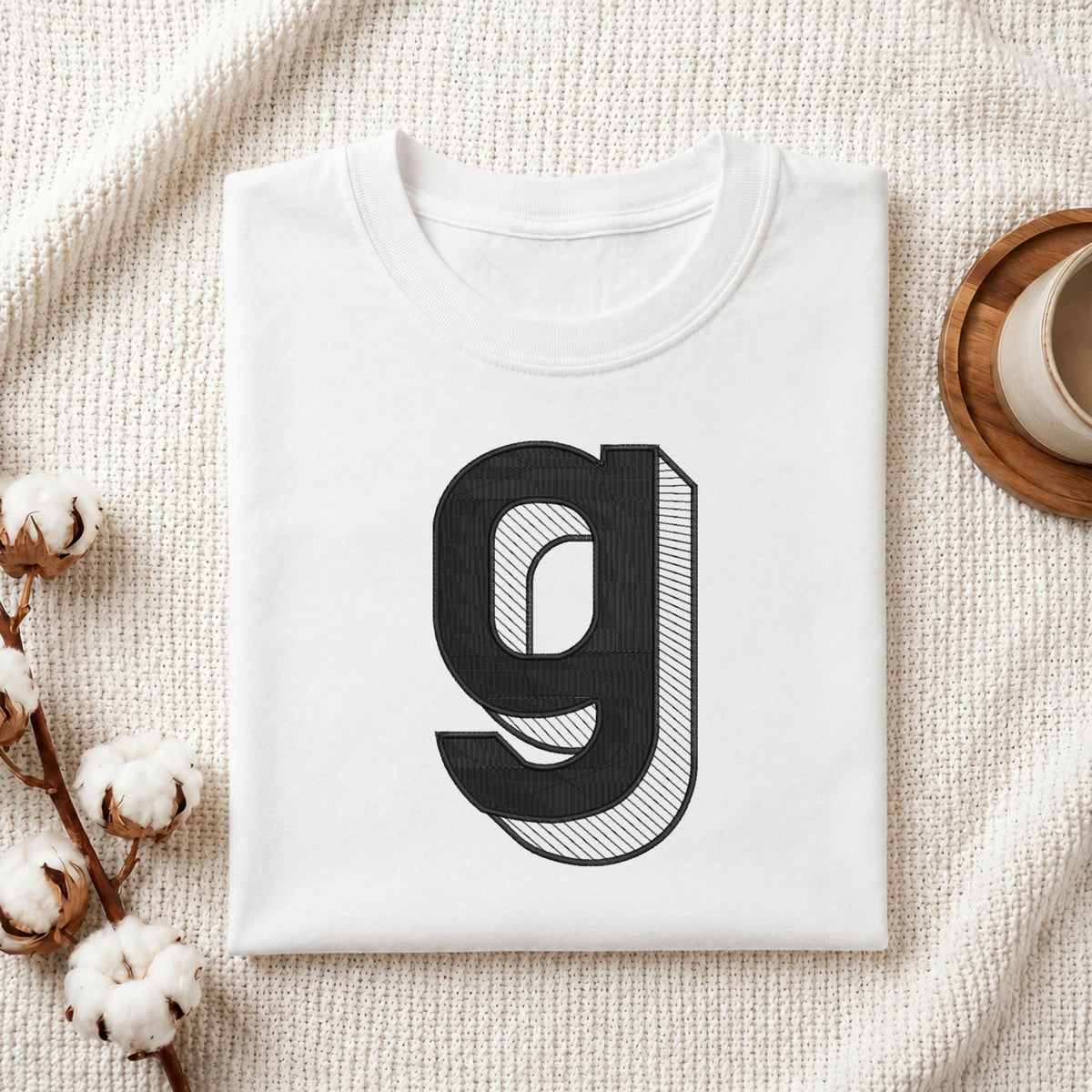 A folded white t-shirt with a large black stylized letter 'g' printed on the front, styled on a textured cream knit blanket with cotton bolls and a wooden cup in the background.
