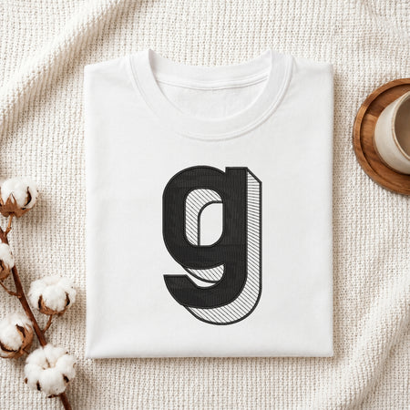 A folded white t-shirt with a large black stylized letter 'g' printed on the front, styled on a textured cream knit blanket with cotton bolls and a wooden cup in the background.