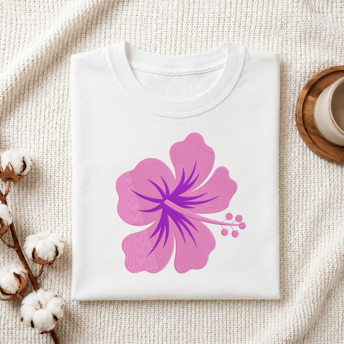 A folded white crewneck t-shirt with a large pink and purple hibiscus flower graphic on the front, styled on a textured cream knit blanket with cotton bolls and a wooden cup in the background.