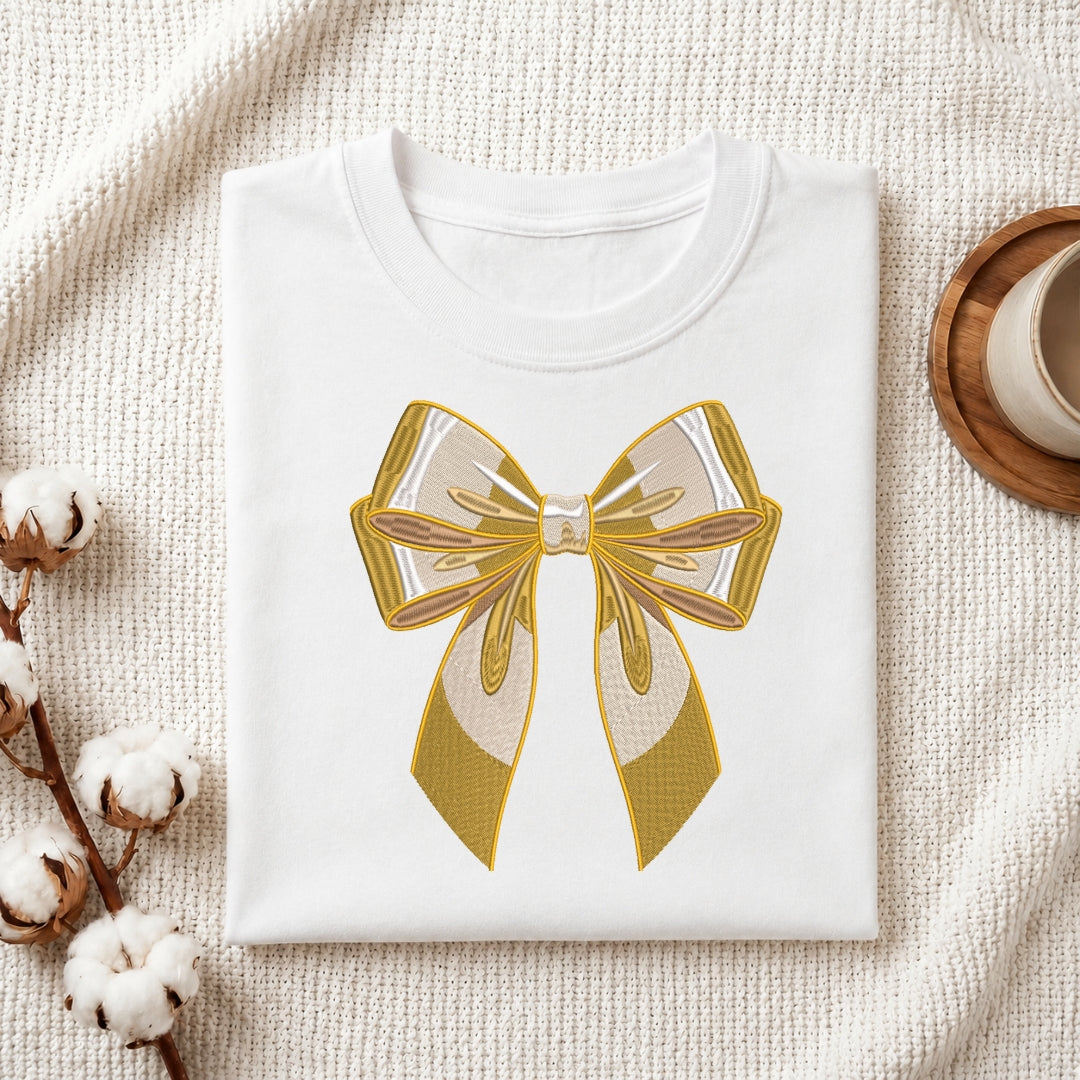 A folded white crewneck t-shirt featuring a large gold and cream bow graphic on the front, styled on a textured beige fabric background with cotton bolls and a wooden cup.