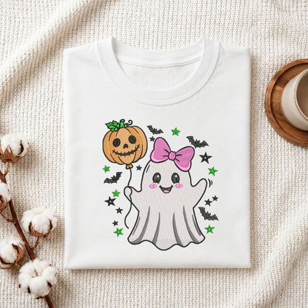 A folded white t-shirt with a cute Halloween graphic, displayed on a textured cream fabric background with cotton bolls and a wooden cup as props. The graphic features a smiling white ghost holding an orange jack-o'-lantern balloon with a pink bow, surrounded by black bats and green stars.