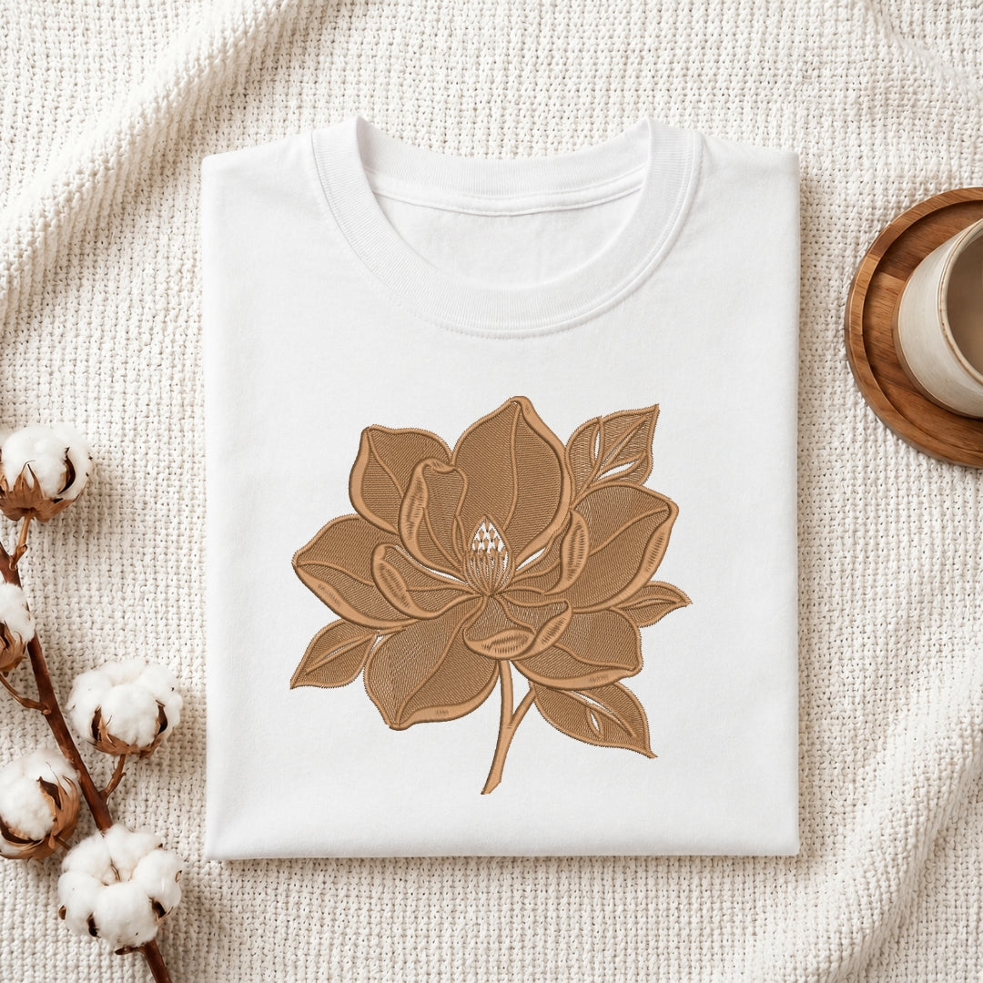 A white folded t-shirt featuring a detailed, brown-toned embroidered flower design, presented on a textured white fabric with cotton sprigs and a wooden coaster.