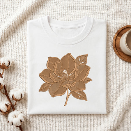 A white folded t-shirt featuring a detailed, brown-toned embroidered flower design, presented on a textured white fabric with cotton sprigs and a wooden coaster.