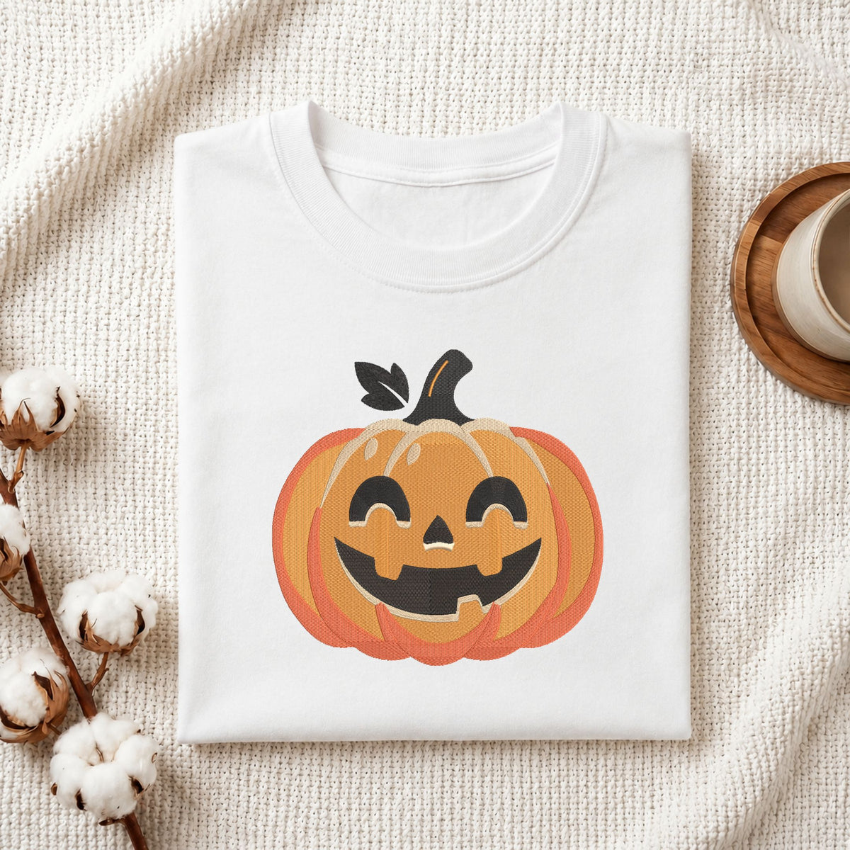 Folded white cotton t-shirt mockup featuring a jack-o'-lantern pumpkin machine embroidery design, styled with cotton stems and a wooden cup.