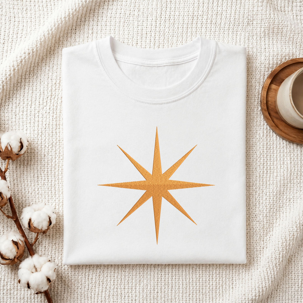 A folded white crewneck t-shirt with a glittery gold 12-point starburst graphic on the front, styled on a textured cream knit blanket with cotton bolls and a wooden cup in the background.