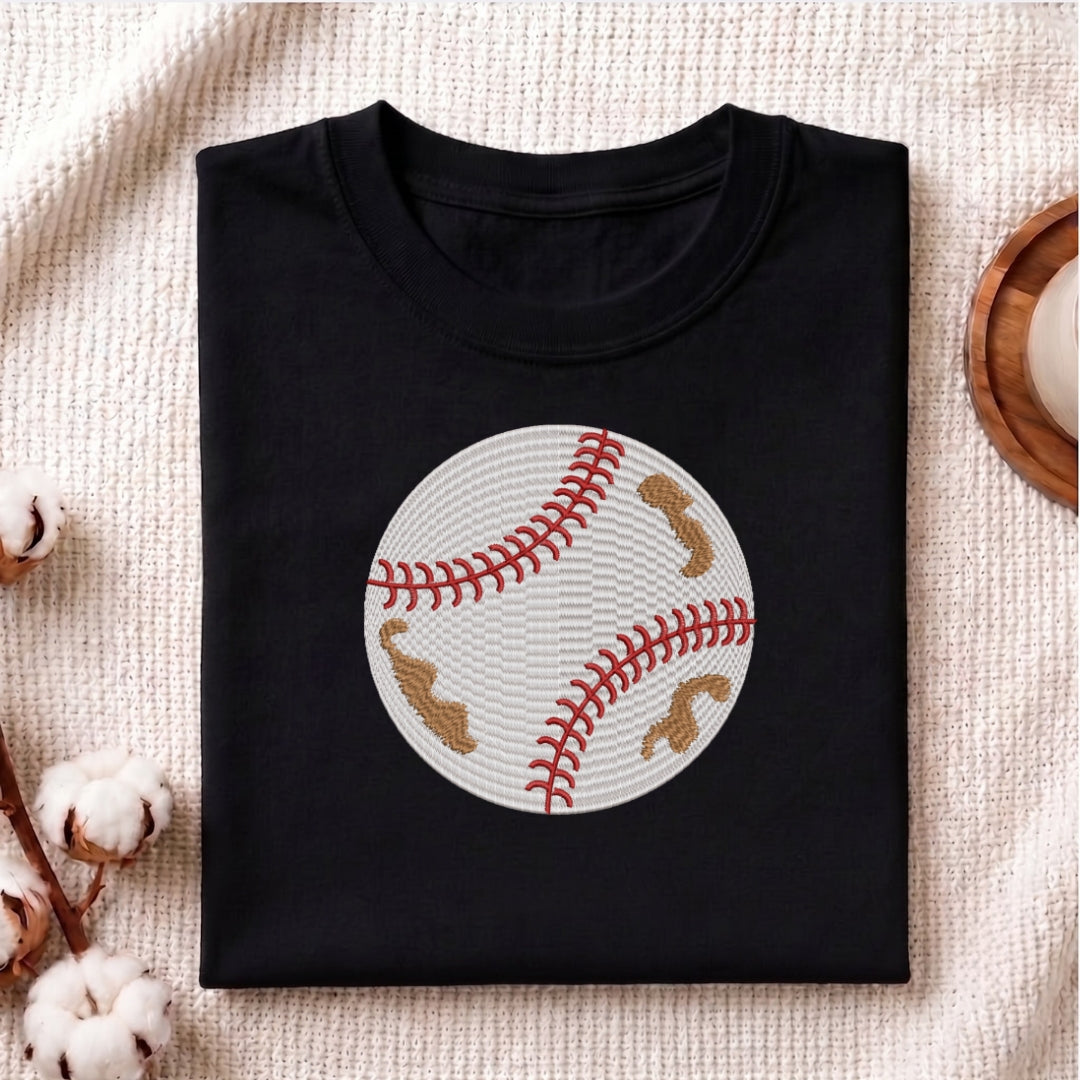 A folded black crewneck t-shirt featuring a baseball graphic with red stitching and tan patches, displayed on a textured beige fabric background with decorative cotton bolls.