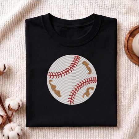 A folded black crewneck t-shirt featuring a baseball graphic with red stitching and tan patches, displayed on a textured beige fabric background with decorative cotton bolls.