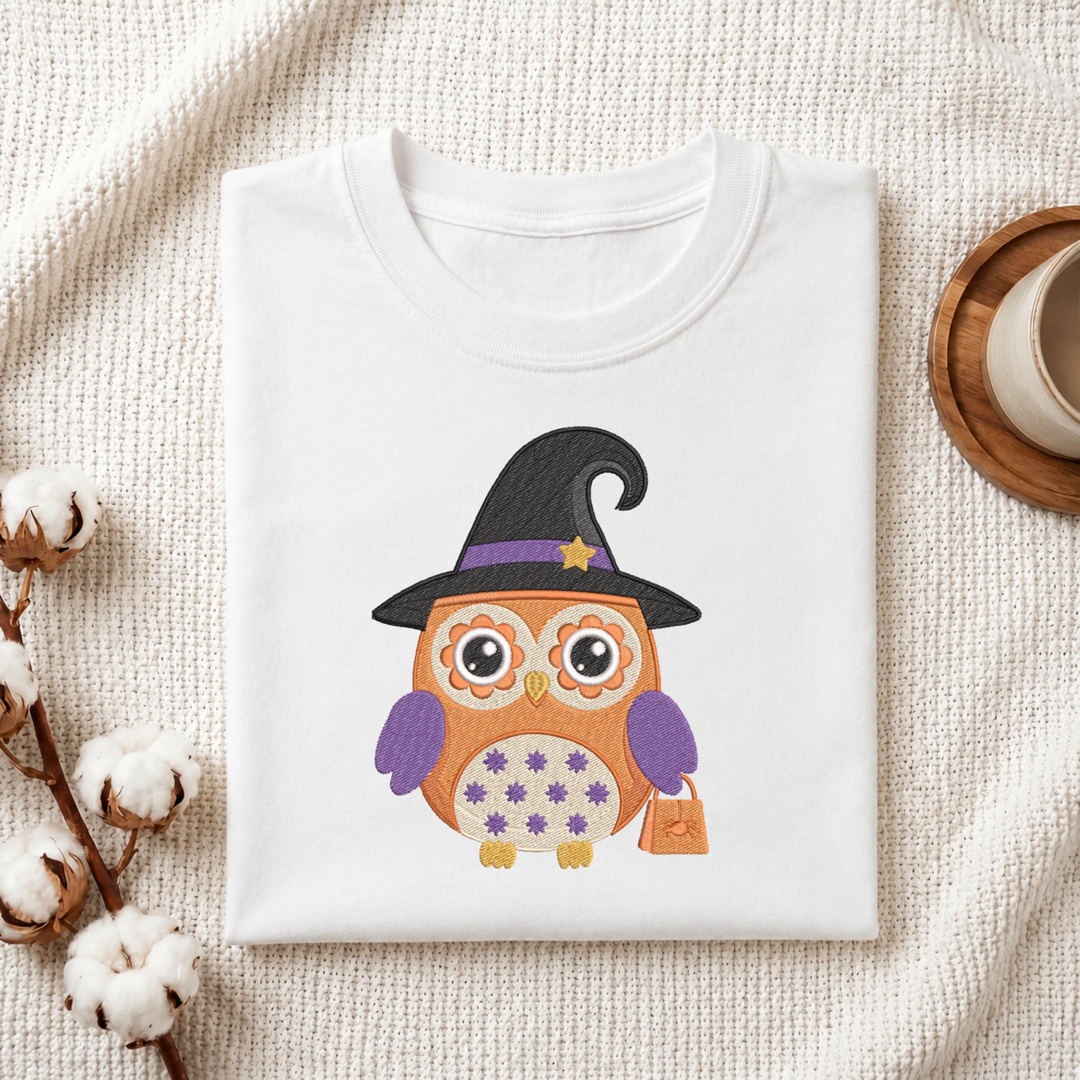 A folded white T-shirt with a colorful embroidered-style owl graphic wearing a black witch hat and purple wings, styled on a textured cream fabric with cotton bolls and a cup in the background.