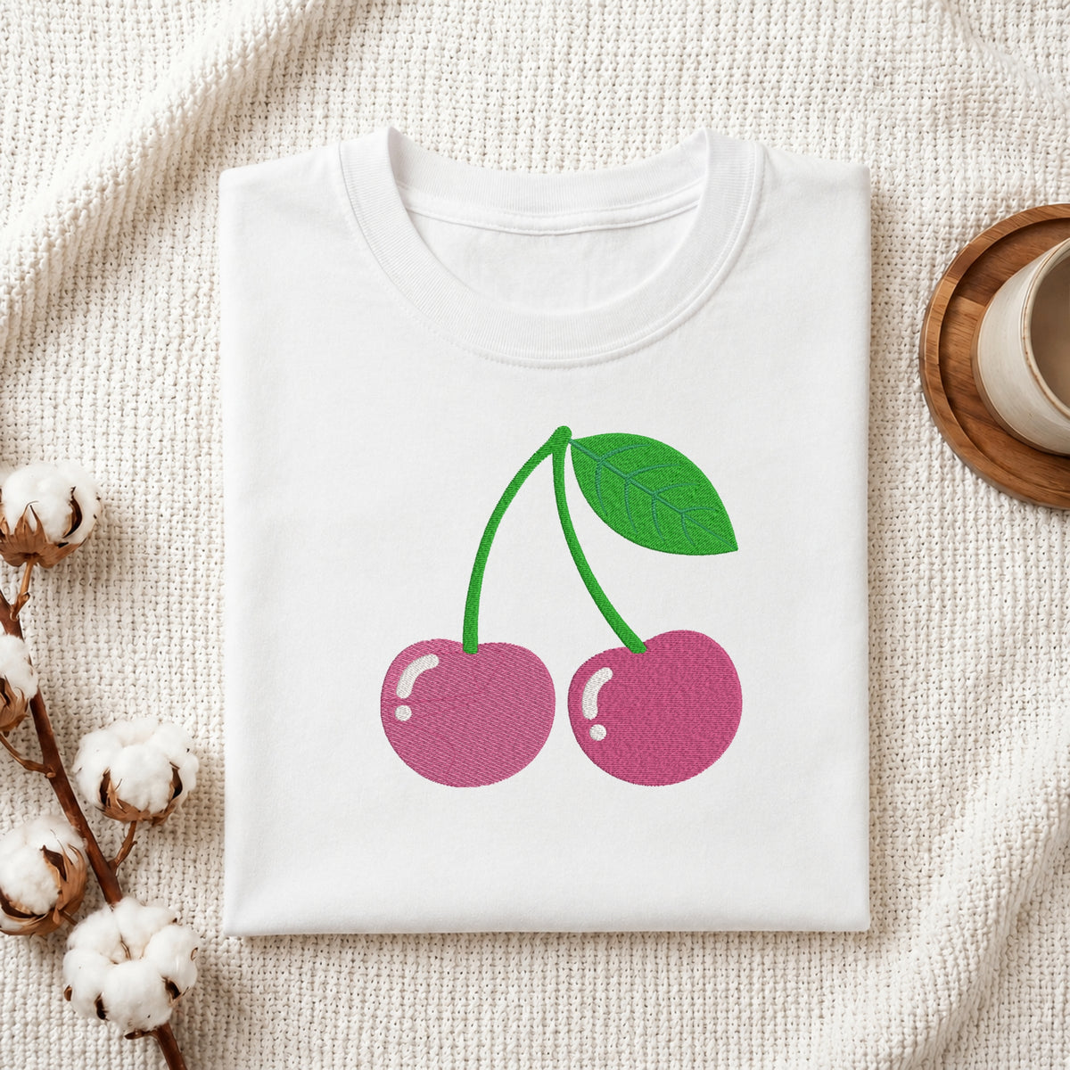 A folded white crewneck t-shirt featuring a graphic of two pink cherries with green stems and a leaf, displayed on a textured cream fabric with cotton bolls and a wooden cup in the background.