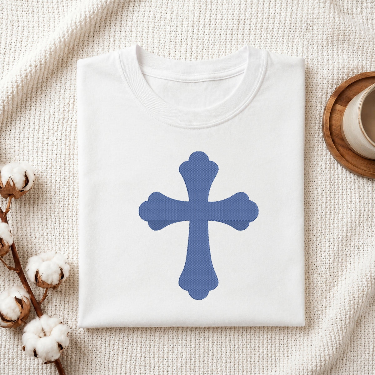 A folded white T-shirt with a blue cross graphic on the front, styled on a textured cream fabric with cotton stems and a small cup in the corners.