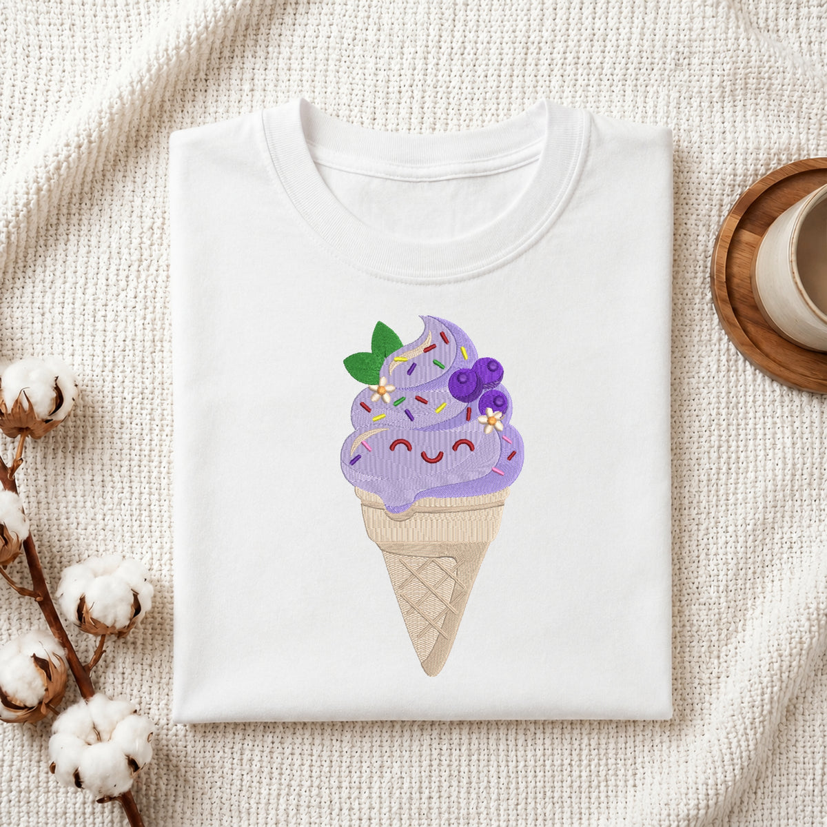A folded white T-shirt with a cute embroidered ice cream cone graphic on the front, styled on a textured cream fabric with cotton bolls and a wooden cup in the background.