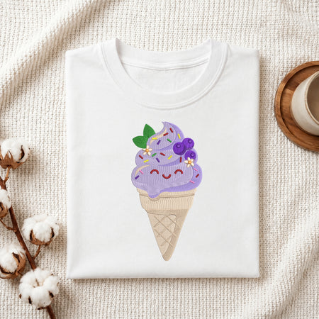 A folded white T-shirt with a cute embroidered ice cream cone graphic on the front, styled on a textured cream fabric with cotton bolls and a wooden cup in the background.