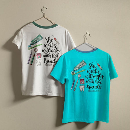 Two short-sleeve ringer T-shirts hanging on hangers against a plain beige wall. The left shirt is white with a teal collar and back graphic featuring dental tools and the text 'She works willingly with her hands PROVERBS 34:23'.