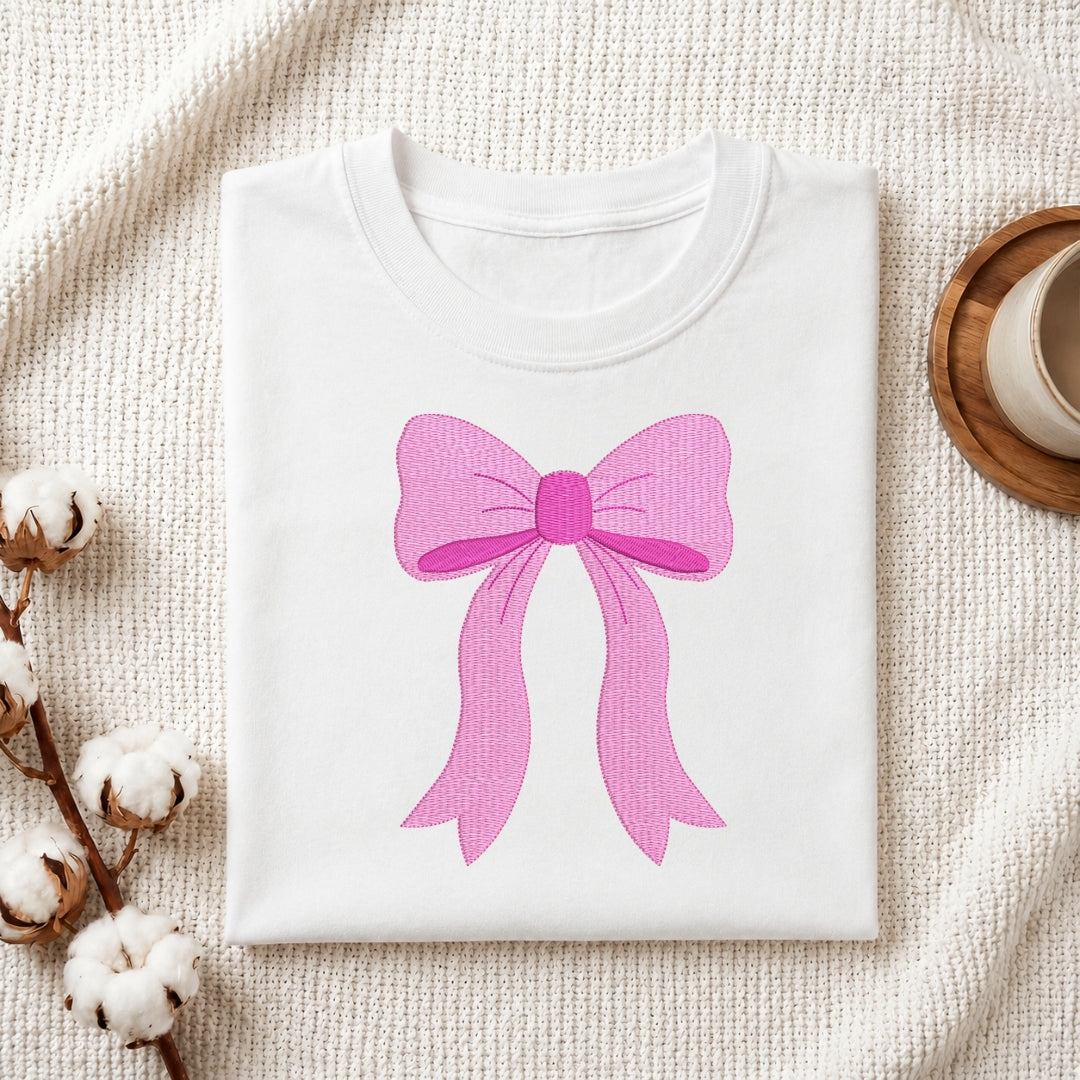 A folded white crewneck t-shirt featuring a large pink bow graphic on the front, styled on a textured cream knit blanket with cotton bolls and a wooden cup in the background.
