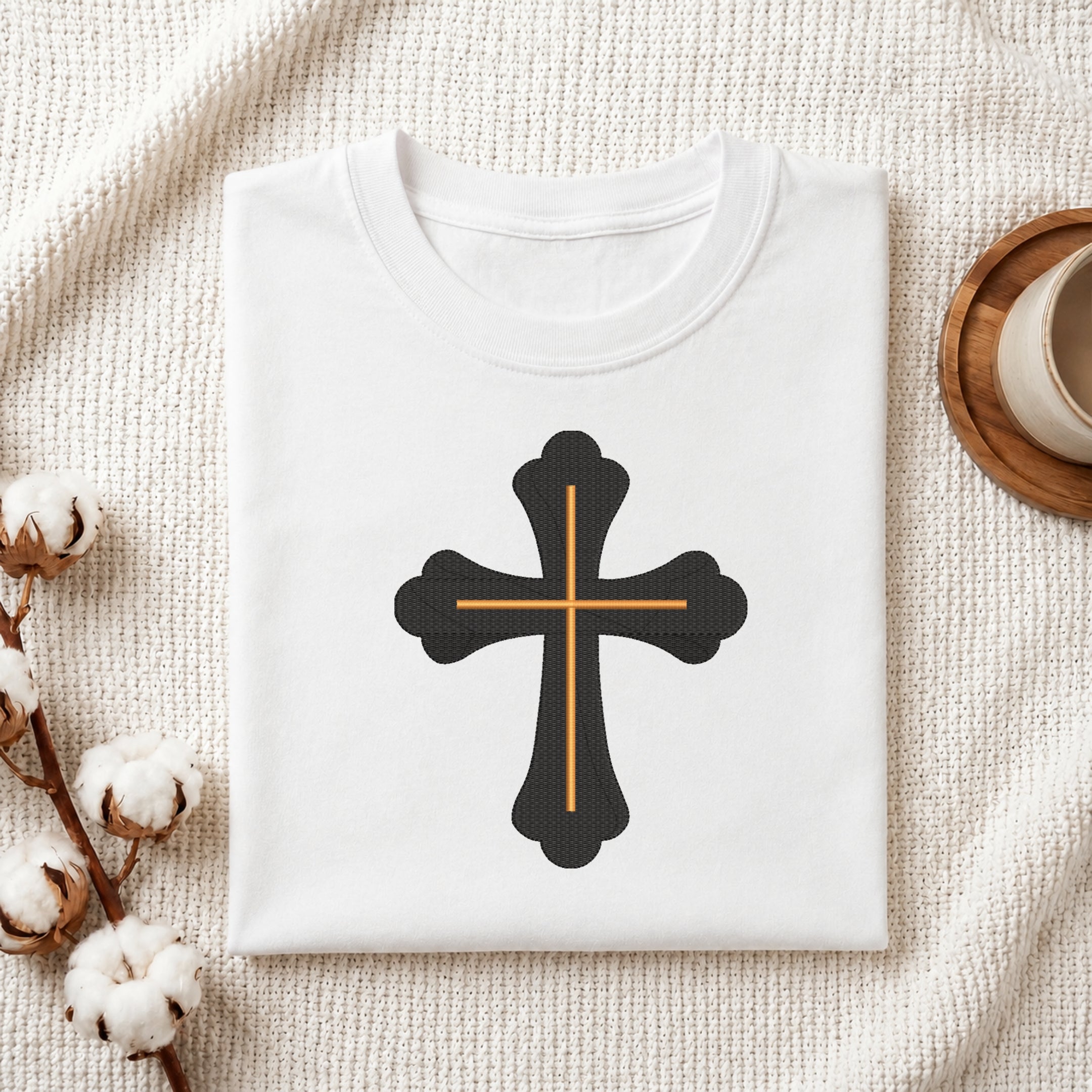 A folded white t-shirt with a black cross graphic outlined in gold, displayed on a textured cream fabric background with decorative cotton bolls and a wooden cup.