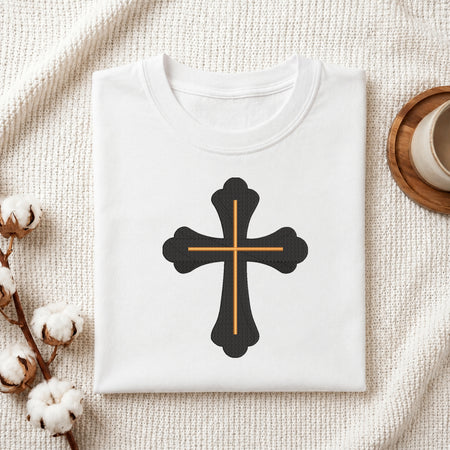 A folded white t-shirt with a black cross graphic outlined in gold, displayed on a textured cream fabric background with decorative cotton bolls and a wooden cup.