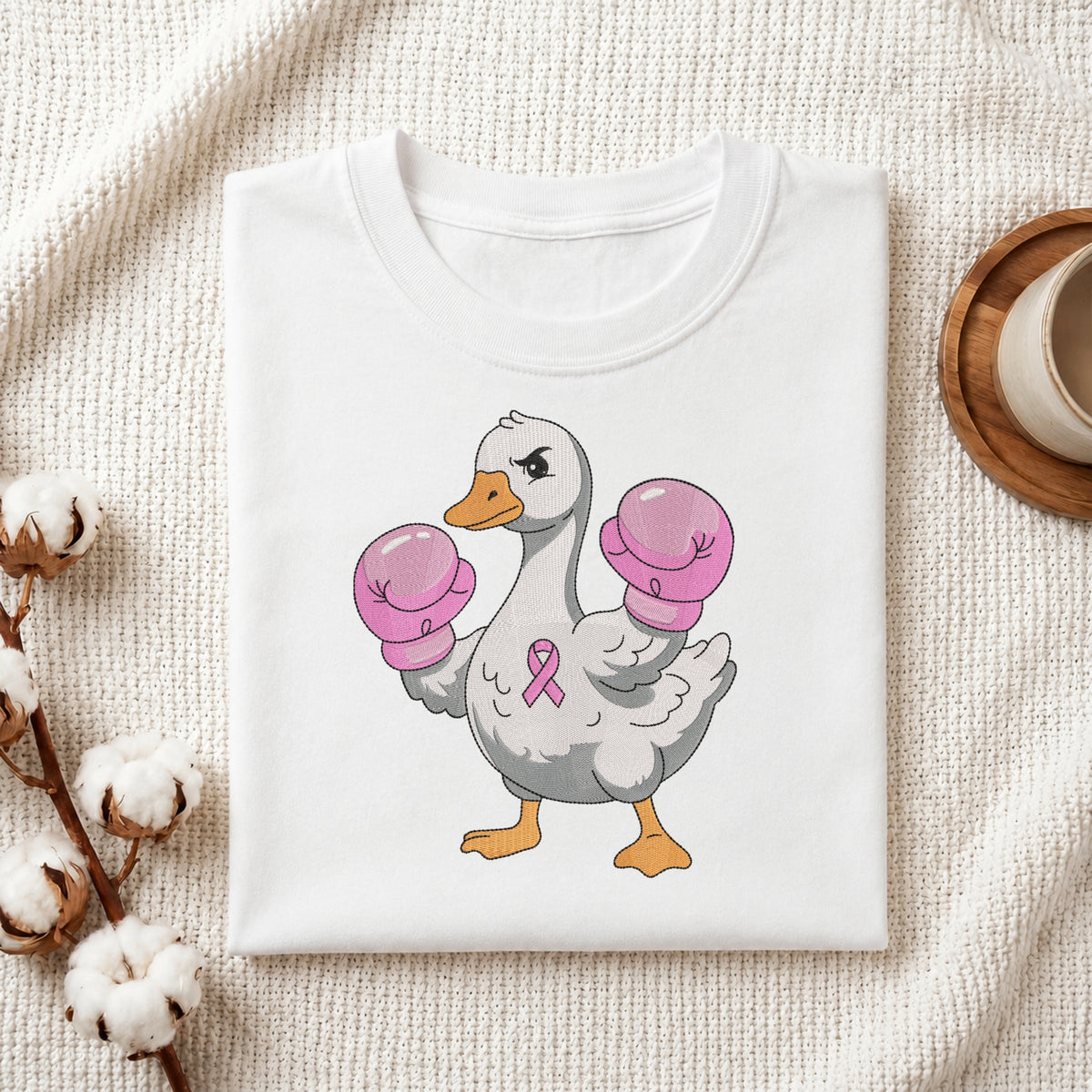 A folded white T-shirt displayed on a textured cream fabric background, featuring a cartoon duck wearing pink boxing gloves and a pink awareness ribbon on the chest. Cotton bolls and a small cup are placed around it as styled props.