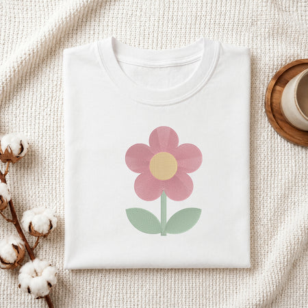 A folded white crewneck t-shirt featuring a retro-style pink flower with a yellow center and green leaves, styled on a textured cream knit blanket with cotton bolls and a wooden cup in the background.