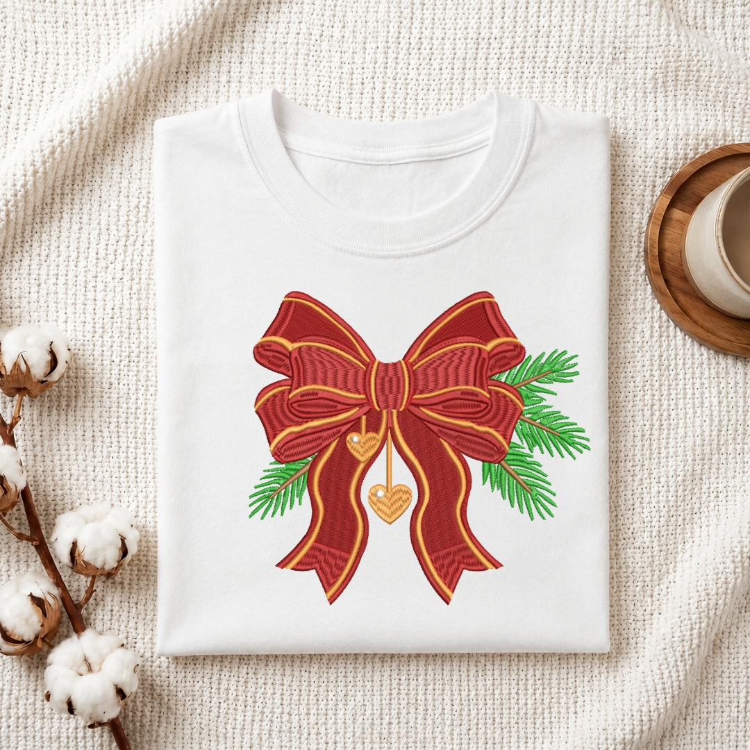 A folded white crewneck t-shirt with a graphic of a red and gold bow, green pine branches, and a gold heart, styled on a textured cream blanket with cotton bolls and a coffee cup.