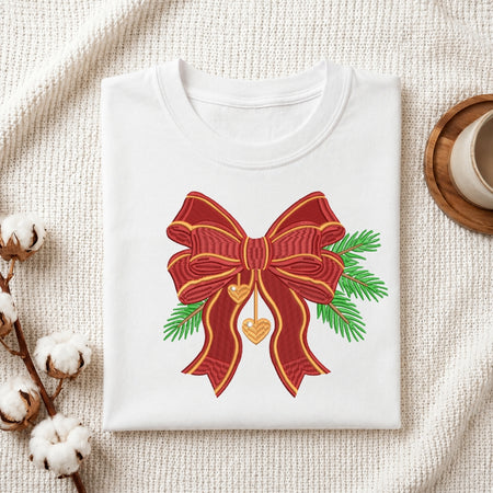 A folded white crewneck t-shirt with a graphic of a red and gold bow, green pine branches, and a gold heart, styled on a textured cream blanket with cotton bolls and a coffee cup.