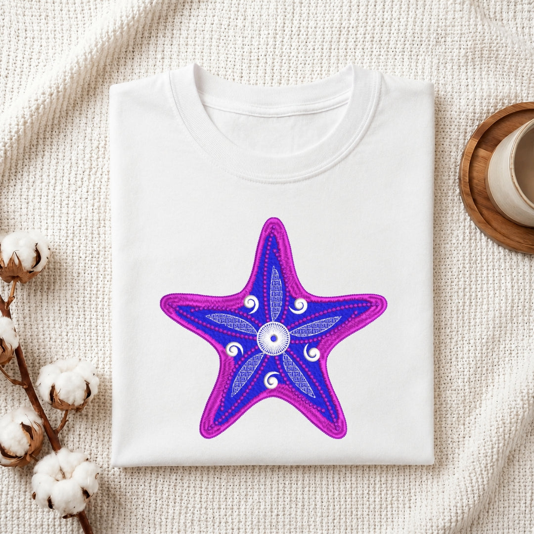 A folded white crewneck t-shirt featuring a vibrant purple and blue starfish graphic with a central white evil eye motif, styled on a textured cream blanket with cotton bolls and a wooden cup in the background.